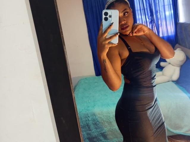 Caylin si fa un selfie allo specchio, indossando un abito nero aderente, senza spalline, che mette in risalto la sua figura sinuosa e atletica. Lei tiene in mano un telefono, appare seducente con delle tende blu sullo sfondo..