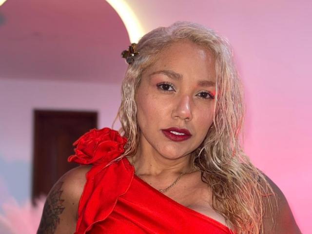 Fotografía de Sonia Ferrer con el cabello rubio y mojado, vistiendo un vestido rojo de un solo hombro y con una gran rosa roja. Ella tiene lápiz labial oscuro, tatuajes en el brazo y una expresión seria. Muy atractivo..