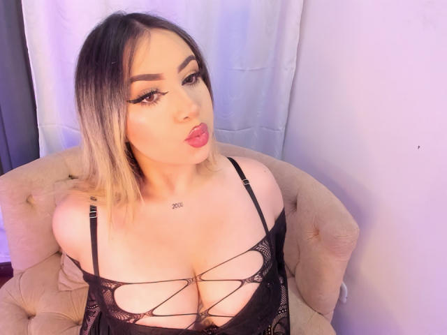 MelissaLust, een Latina vrouw met blond haar dat tot op haar schouders reikt, volle rode lippen en veel eyeliner, zit op een beige gestoffeerde stoel. Ze draagt een zwarte top met een open, ruitvormig patroon, waardoor haar grote borsten zichtbaar zijn. Helder witte achtergrond.