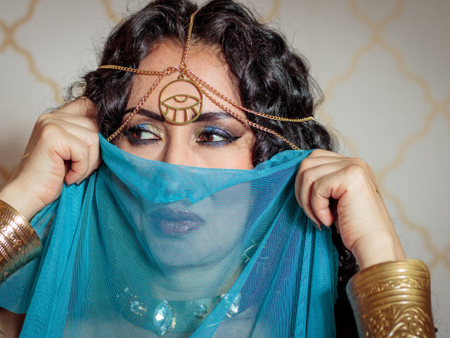 Samantha Antolín, cabello oscuro y ondulado, tocado dorado con diseño circular, sosteniendo un velo de malla azul sobre el rostro, sombra de ojos azul. pulseras doradas en las muñecas, mirada intensa, tensión sexual, colores vibrantes, texturas intrincadas..