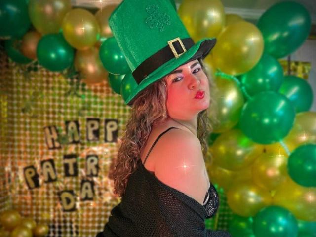 AdrianaPrettie, met krullend bruin haar, draagt een groene, glinsterende hoed en een zwarte top met een open schouder, omringd door groen. en gele ballonnen. "Vrolijke Patti-dag" bordje op de achtergrond. Ze kijkt om, met een frons..