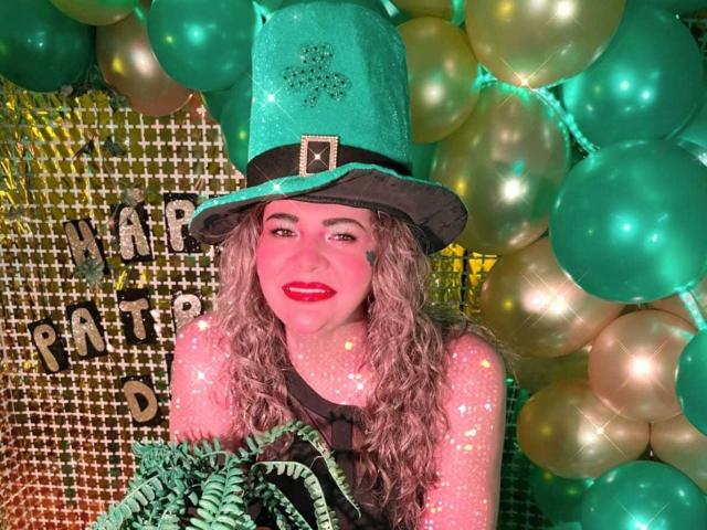 AdrianaPrettie mit welligem, blondem Haar, leuchtendem rotem Lippenstift und einem grünen, glitzernden Zylinder, umgeben von Türkis und Gold. Ballons. Ihre Hand ruht auf ihrer Wange, und sie lächelt verführerisch..