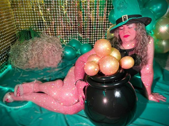 AdrianaPrettie, con cabello castaño largo y rizado, usando un sombrero de bruja verde, un collar negro y un conjunto rosa brillante, recostada. sobre una tela verde, rodeado de globos dorados brillantes y una olla negra..