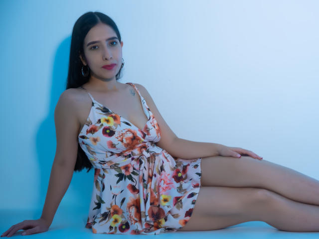 Ivana Clark está sentada contra una pared iluminada con un color azul brillante, con las piernas extendidas, vestida con un vestido de verano floral con un escote en V pronunciado, que muestra su figura delgada y sus pequeños senos. Ella mira directamente a la cámara con una expresión neutra.