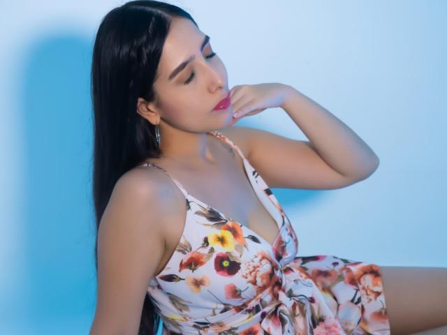 Ivana Clark, una mujer latina con cabello largo y negro, lleva un vestido floral con un escote bajo que deja ver parte de su pecho. Está posando de forma seductora frente a un fondo azul, con la mano cerca de la boca. Increíblemente atractivo/a..