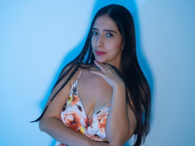 Fotografia de Ivana Clark, em pé em frente a uma parede iluminada em azul, vestindo um vestido floral com decote profundo, cabelos pretos, mão em close, expressão séria, a iluminação azul realça sua figura, sensualidade e tensão..