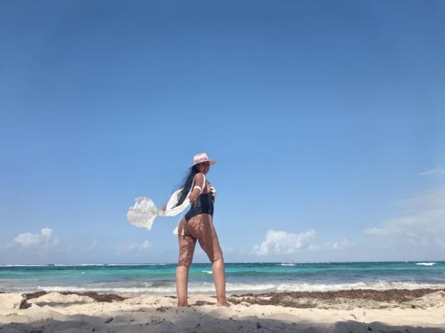 JackieMature stands on a sandy beach, facing the ocean. She's wearing a black swimsuit and white cap, her long black hair flowing in the wind. Bright blue sky, turquoise water. Fucking hot.