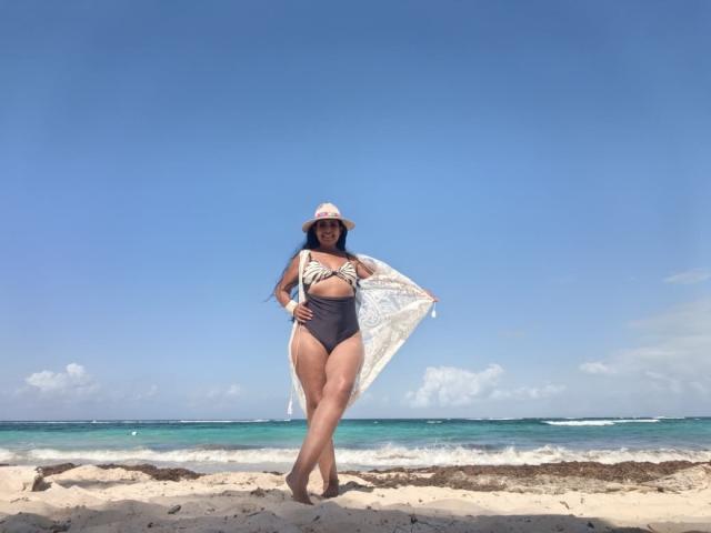 Jackie Mature, eine kurvige Frau mit heller Haut, steht an einem sandigen Strand. Sie trägt einen schwarzen Badeanzug und hält einen weißen Fächer aus Spitze. Ihre Beine sind leicht gespreizt, was Selbstbewusstsein zeigt. Im Hintergrund ist ein klarer, blauer Himmel und das Meer zu sehen..