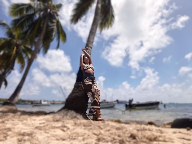 JackieMature está em uma praia de areia, encostada em uma palmeira, vestindo calças e botas de camuflagem, com uma arma apontada. mãos no cabelo, barcos ao fundo, céu azul com nuvens.