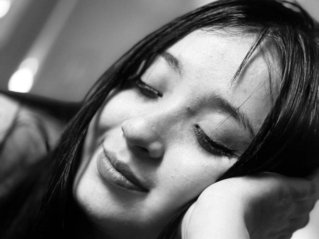 Schwarzes und weißes Nahaufnahmefoto von LunaBellaHotty mit langen, dunklen Haaren, geschlossenen Augen und einem sanften Lächeln. Sie legt ihren Kopf auf ihren Arm und erzeugt so eine sinnliche, entspannte Atmosphäre..