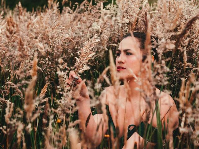 Elizabeth Dupont, con cabello oscuro, en un campo de hierba alta y beige. Ella sostiene una flor, con los hombros desnudos a la vista, y tiene una expresión concentrada, ligeramente sensual. Tensión erótica en un entorno natural..