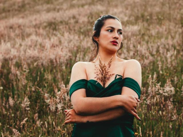 Elizabeth Dupont staat in een veld met hoog, rozeachtig gras, met haar armen over elkaar geslagen. Ze draagt een groene jurk met een open schouder, en haar donkere haar is naar achteren getrokken. Haar uitdrukking is intens, aantrekkelijk..