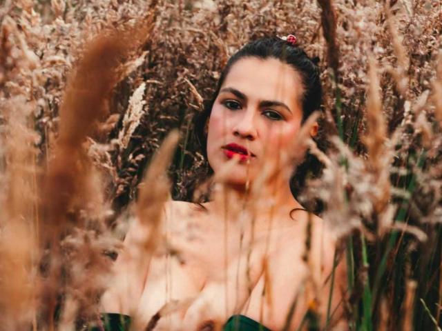 Fotografie von Elizabeth Dupont mit dunklem Haar in einem Feld mit hohem, braunem Gras. Sie trägt roten Lippenstift und ein trägerloses Oberteil und blickt direkt in die Kamera mit einem verführerischen, sinnlichen Ausdruck. Unschärfer Gras im Vordergrund.