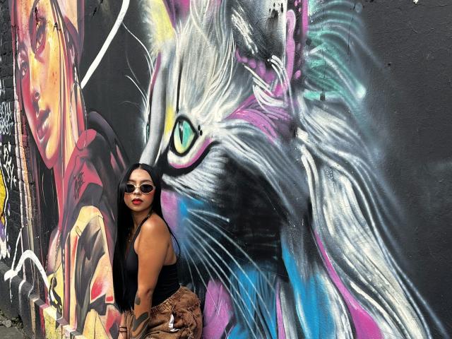 ChloeMoretti se tient devant une immense fresque de graffiti, colorée, représentant un chat blanc lumineux aux yeux néon. Elle porte un haut noir et des lunettes de soleil, et regarde directement l'appareil photo. Ambiance extrêmement intense..