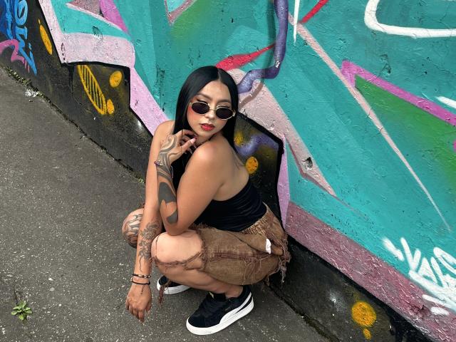 Chloe Moretti est accroupie dans une rue recouverte de graffitis, vêtue d'un haut noir, d'un short beige et de baskets noires. Elle a de longs cheveux noirs, du rouge à lèvres, des lunettes de soleil et un tatouage sur son bras gauche. Extrêmement séduisant..