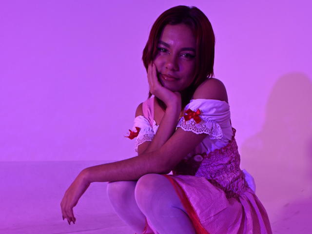 NikkyDoll squats in front of a purple-lit background, wearing a white off-shoulder dress with red flowers and white leggings. She rests her face on her hand, smiling slightly. Bright purple light highlights her brown hair.