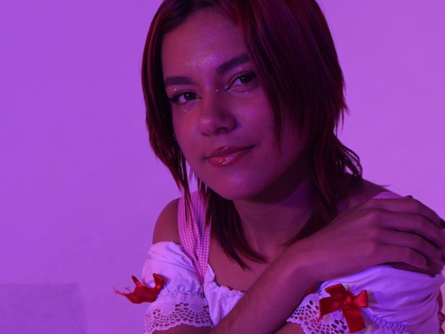 Close-up photo of NikkyDoll with medium brown skin, brown hair, and subtle makeup. She's wearing a white off-shoulder top with red bows. Purple backlight highlights her shiny lips and relaxed, confident expression.