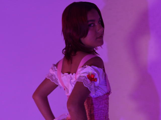 Photograph of NikkyDoll with shoulder-length brown hair, wearing a white off-shoulder dress with red flowers, back to camera, purple and pink lighting, shadow on right, suggestive pose, sexual tension.