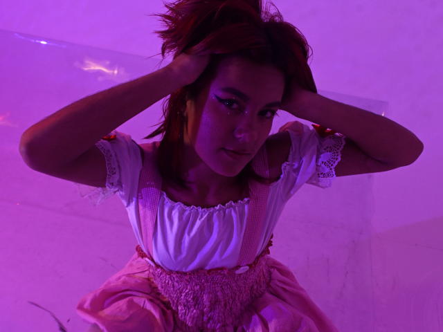 NikkyDoll met donkere huid en rood haar, gekleed in een witte blouse en een roze jurk, handen in haar haar, intense blik, paars en roze verlichting, verleidelijke pose, zichtbare spanning, sensuele sfeer.