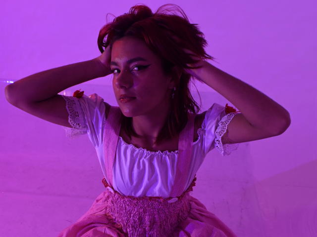 NikkyDoll, avec ses cheveux foncés, les mains dans les cheveux, portant une blouse blanche avec des manches en dentelle et un tablier rose à carreaux, baignée dans lumière violette.
regard séduisant et intense, visage ombragé, texture douce du tissu..
