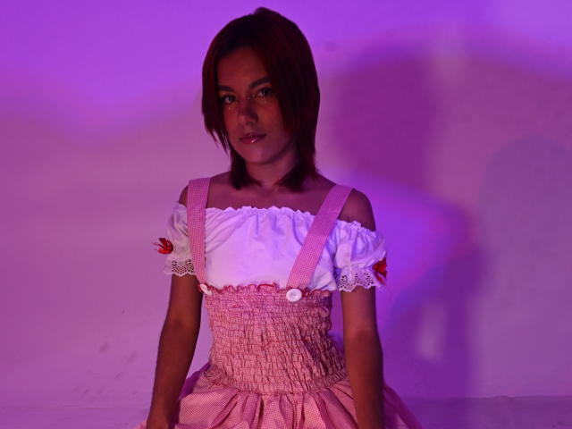 NikkyDoll sitzt mit gespreizten Beinen, trägt ein rosa und weißes Kleid mit einem Schulterteil, das herunterfällt, mit Puffärmeln und roten Knöpfen. Das violette und rosa Licht wirft einen sinnlichen Schein auf ihr Gesicht..