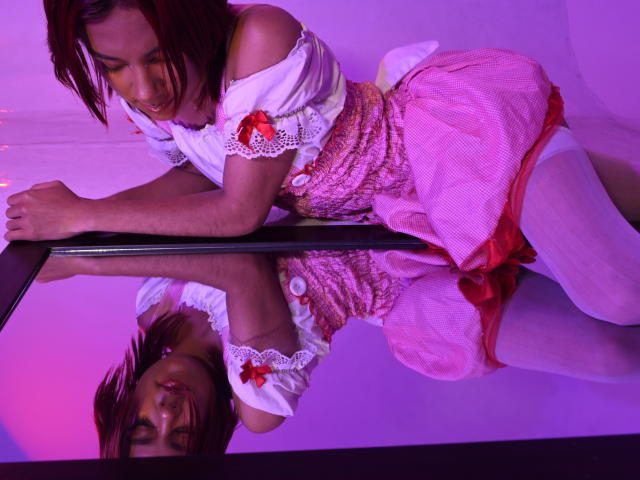 NikkyDoll è appoggiata a una superficie riflettente, indossa una maglietta bianca con spalline scoperte con fiocchi rossi, una gonna a quadretti rosa e calze bianche.
l'illuminazione rosa acceso e viola esalta la sua posa seducente..