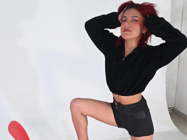 Photograph of NikkyDoll, posing with hands in hair, black cropped jacket, black shorts, one leg raised, red object on floor, white background, red highlights in hair, confident expression.