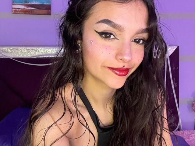 Close-up photo of MarietteJones, fair-skinned woman with long black hair, red lipstick, black eyeliner, nose piercing, black halter top, smiling suggestively, blue and purple lighting, bedroom background. Fucking hot.