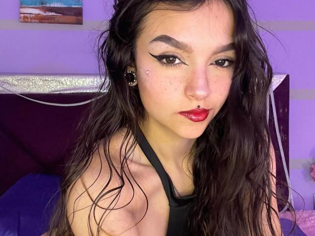 Close-up van Mariette Jones, met bleke huid en donker, golvend haar, die een zwarte tanktop draagt. Ze heeft opvallende, zwarte eyeliner, rode lippenstift en een neusring. De achtergrond heeft paarse en blauwe verlichting..