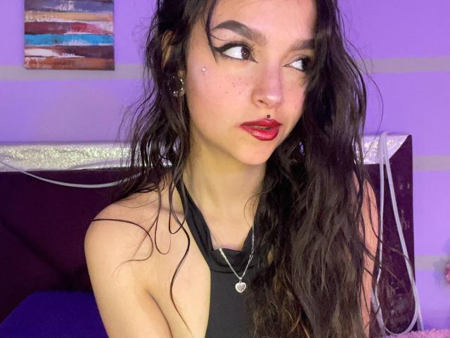 Fotografia di Mariette Jones, una donna dalla pelle chiara con lunghi capelli neri bagnati, rossetto rosso e eyeliner scuro, che guarda. di lato.
indossa un top nero con spalline, appare sensuale, con un sorriso appena accennato..