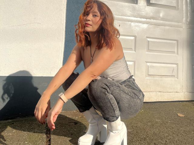 Photographie d'Emily Klein accroupie contre une porte blanche, vêtue d'un débardeur gris, d'un jean foncé et de bottes blanches. Ses cheveux roux sont en désordre, créant une ambiance sexy et intense. La lumière du soleil met en valeur ses courbes..