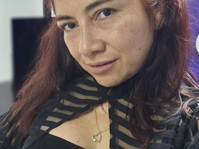 Fotografia ravvicinata di AlessiaRed, una donna con la pelle color nocciola e i capelli rossi, che indossa una maglia nera trasparente con diagonali. righe che lasciavano intravedere il décolleté e una collana d'oro. Ha un leggero sorriso, guarda direttamente la fotocamera con un'espressione sottile e seducente..