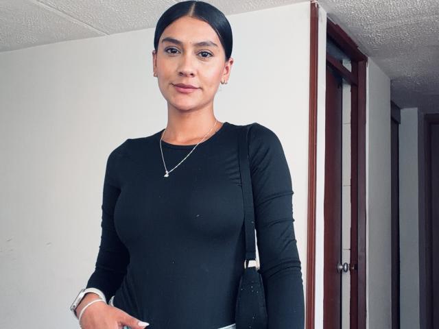 ThaliaAres, a young woman with medium brown skin and black hair in a tight bun, wearing a tight black long-sleeve top, standing in a doorway, looking directly at the camera.