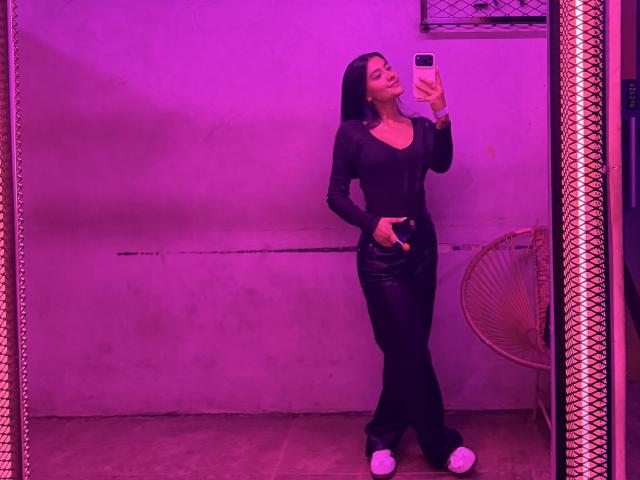 ThaliaAres, con i capelli scuri e un abito nero, è in una stanza illuminata di rosa e tiene in mano un telefono per scattare un selfie allo specchio. La sua mano nei pantaloni, l'erezione visibile, una tensione sensuale..