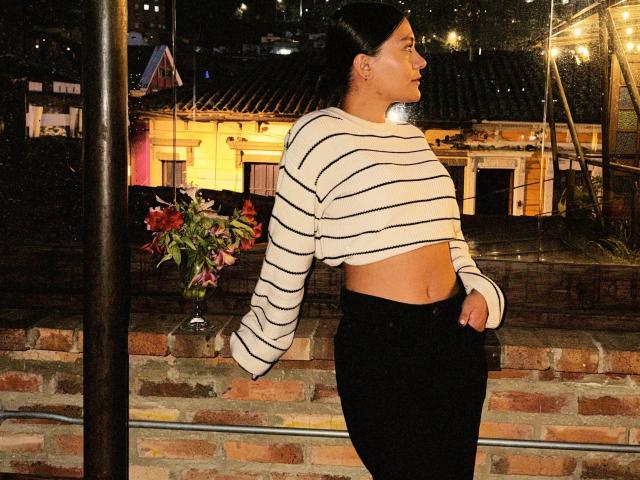 Thalia Ares está do lado de fora, à noite, encostada em uma parede de tijolos. Ela veste uma blusa cropped branca com listras e mangas compridas, e calças pretas. A mão dela repousa no quadril, e ela olha para o lado. Luzes quentes brilham ao fundo..