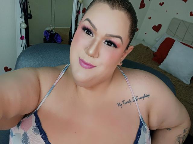 Fotografie einer kurvigen Frau mit heller Haut, dunklem Haar, rosa Lippenstift und schwarzem Eyeliner, die ein gemustertes oberteil mit Spaghetti-Trägern, das ein Tattoo an ihrer rechten Schulter zeigt. Sie schaut mit einem leichten Lächeln in die Kamera. Ihre Brüste sind groß, und ihre Brustpartie ist deutlich sichtbar. Rote Herzformen sind im Hintergrund zu sehen..