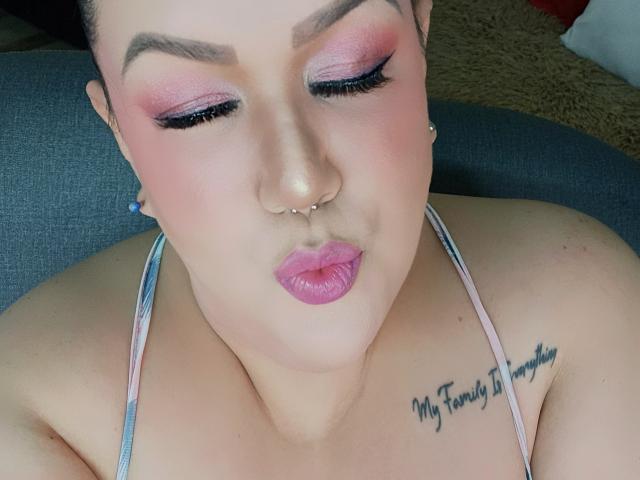 Close-up of GiiannaMorrys with pink eyeshadow, black eyeliner, and pink lipstick. She has a nose piercing and a tattoo on her shoulder reading "My Family Ties My Hands." She's looking down with a slightly pouty expression.