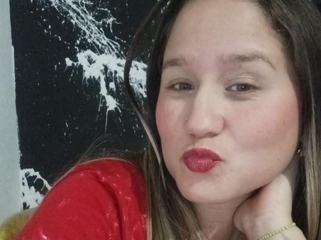Close-up of AlexaHotSeins, light-skinned woman with brown hair, red lipstick, and red shirt, pouting with hand on neck. Background has black and white abstract splatters. Fucking sexy expression.