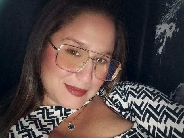 Close-up of AlexaHotSeins, wearing large rectangular glasses, black-and-white patterned top, red lipstick, and a blue pendant. She leans forward, slightly smiling, with visible sexual tension. Dark background.