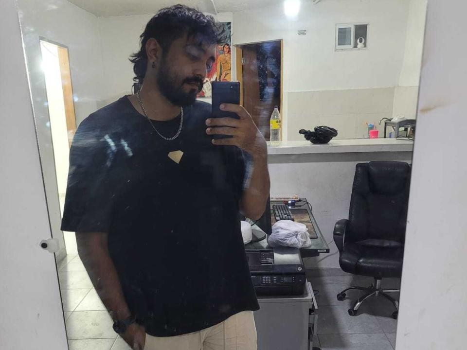 LatinBouser, um homem de pele escura com barba e cabelo despenteado, aparece em uma selfie em frente a um espelho, vestindo uma camiseta preta e calças brancas, segurando um telefone, com uma cadeira e roupas sujas ao fundo..