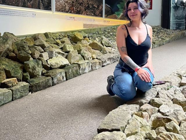 DarckVelvet kneels on a path, wearing a black tank top and blue jeans, with her hands on her knees, surrounded by rough stone blocks. Bright light highlights her curvy figure.
