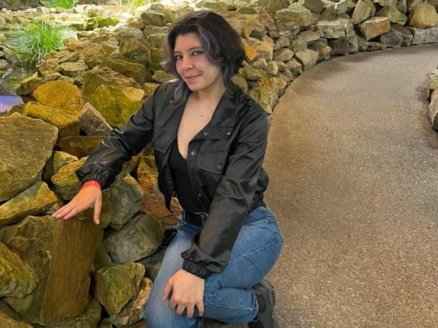 DarckVelvet kneels against a rock wall, wearing a black leather jacket and blue jeans. She has short black hair, smiling, with a slight lean, showing her curvy hips and ass.