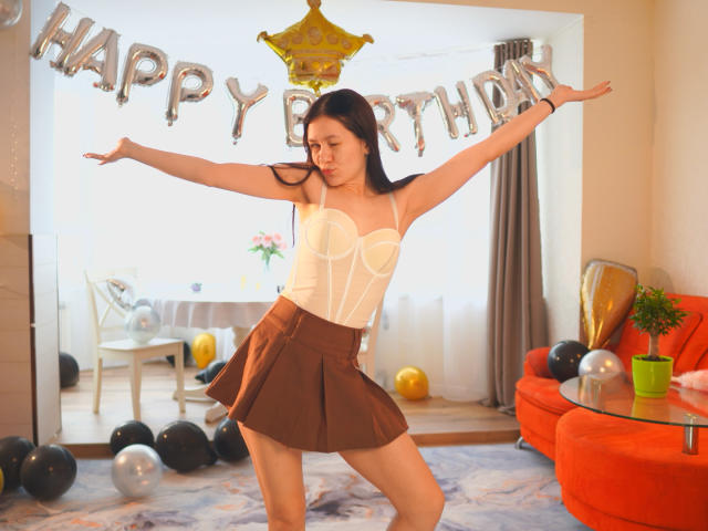 SenaCushingBerry, posant de manière suggestive avec les bras tendus, portant un corset blanc et une jupe marron, dans un décor très éclairé. Pièce avec des ballons "Joyeux anniversaire", un canapé orange et des décorations de fête. Elle est séduisante et sûre d'elle..