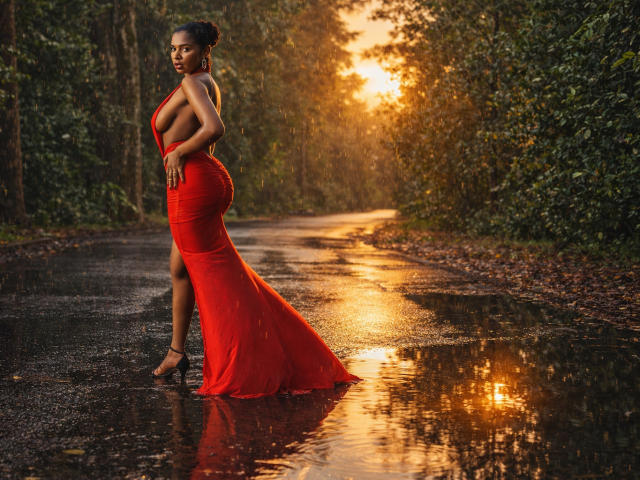 LisaMendez vestida con un vestido rojo ajustado, sin tirantes y con una larga cola, de pie en una carretera mojada al atardecer. La luz del sol resalta sus curvas, su piel oscura y su cabello mojado. Gotas de lluvia en su vestido..