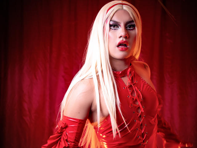 StefyBratz, avec de longs cheveux blonds, un maquillage prononcé et une tenue en latex rouge. Lumière rouge, regard intense, léger sourire. Gants rouges, épaules dénudées. Tension sexuelle, pose provocante, couleurs vives. Éclairage vif et dramatique..
