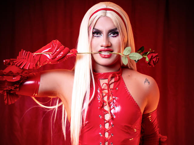 StefyBratz with platinum blonde hair, wearing a shiny red latex corset and matching gloves, holding a rose between her teeth. Intense red lighting, smoky eye makeup, tattoo on left shoulder.