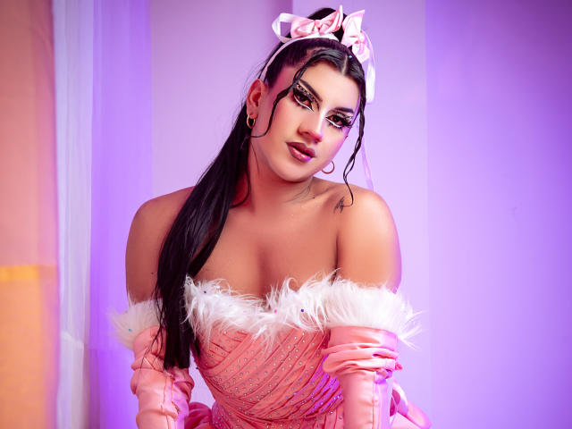 StefyBratz, vestida con un vestido rosa y peludo con un corte asimétrico en el hombro, se inclina hacia adelante con una expresión seductora. Ella tiene el pelo largo y negro, mucho maquillaje y una diadema rosa con un lazo. Iluminación en tonos púrpura y rosa brillantes..