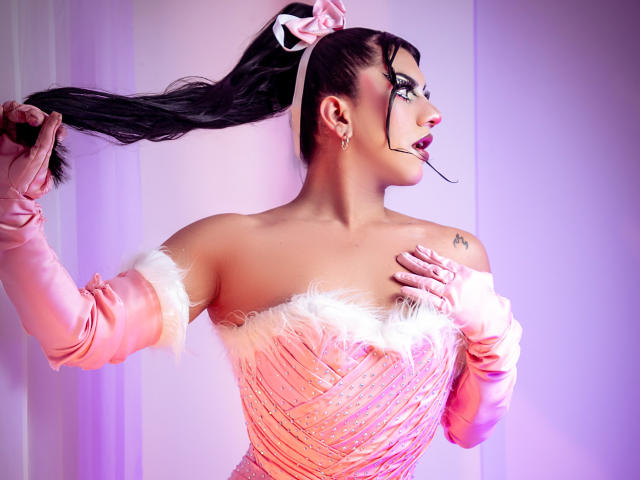 StefyBratz, vestindo um vestido rosa com detalhes de pele e alças que caem do ombro, segura seu cabelo preto com uma mão, enquanto a outra mão está em seu peito, chupando uma pirulito, perfil lateral, iluminação roxa neon.