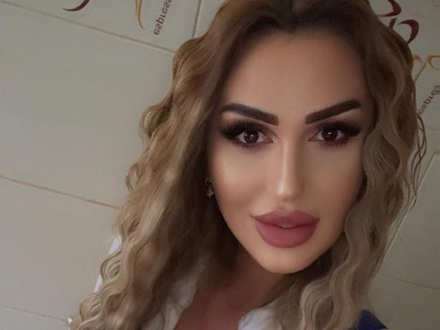 Autoportrait en gros plan de NicolSNaya, avec des cheveux blonds ondulés, un maquillage prononcé, un rouge à lèvres rouge foncé et un fard à paupières fumé. Elle regarde directement l'appareil photo avec une expression légèrement séduisante..