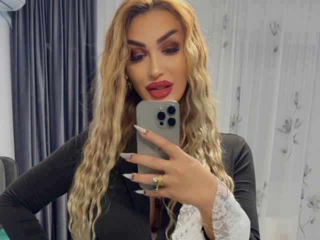 NicolSNaya se toma un selfie frente al espejo, sosteniendo un iPhone gris con tres cámaras. Ella tiene el pelo rubio, largo y ondulado, lápiz labial rojo y cejas pobladas. Usando una blusa negra con mangas de encaje blanco.